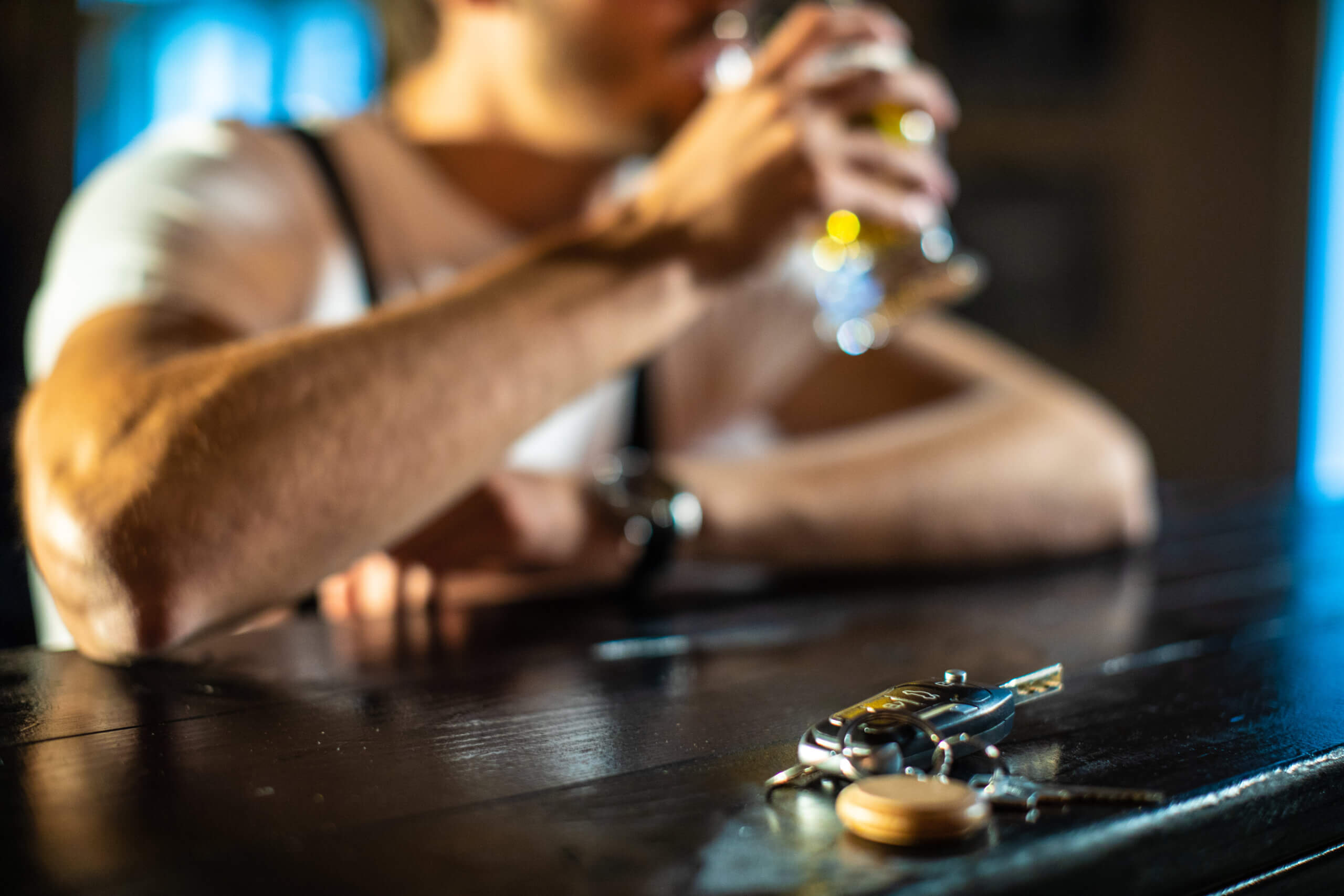 A man sits at a bar, holding a beer in one hand and a key in the other, appearing relaxed and contemplative